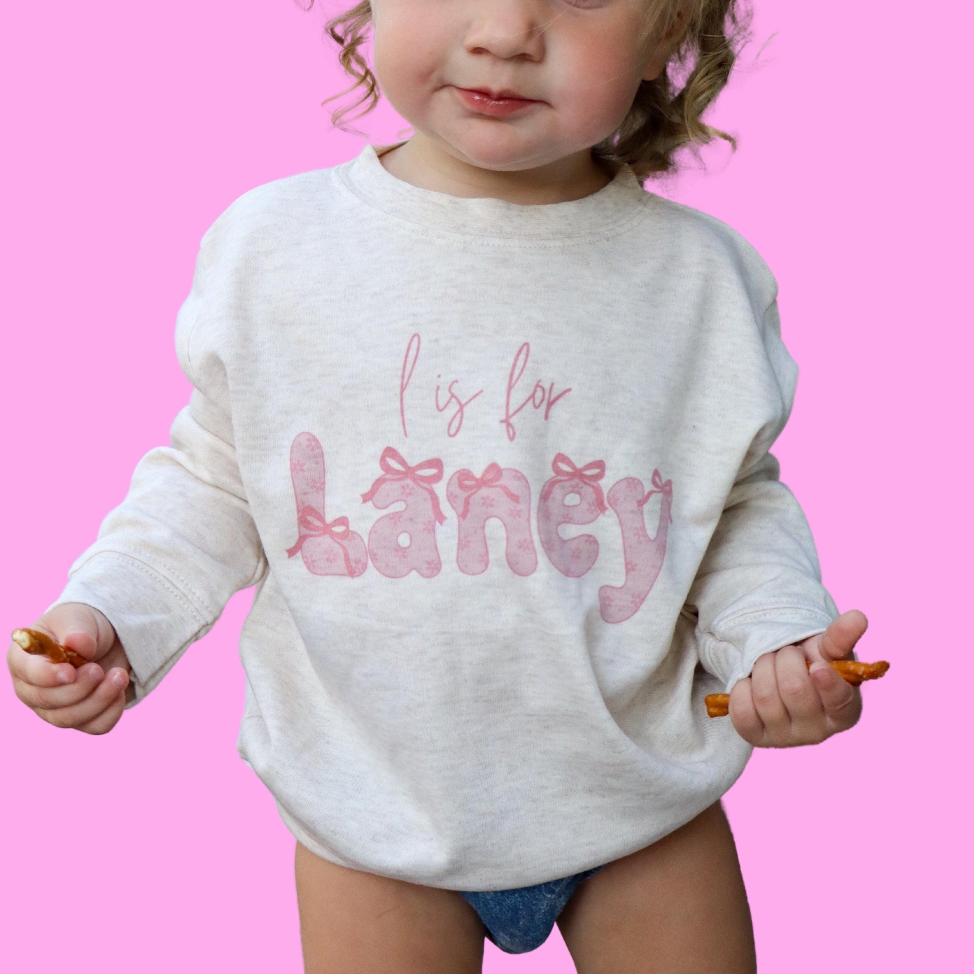 girls custom coquette bow name shirt. pink coquette bow name font printed on a oatmeal colored shirt of your choice.